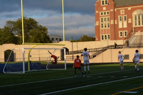 Goalkeeper Jeremiah Higgins in midair jumping for the ball while it shoots past him and into the net.  